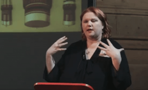 Robyne Calvert giving a talk, gesturing expressively with both hands. She stands behind a podium, with a projected image of lighting fixtures behind her.