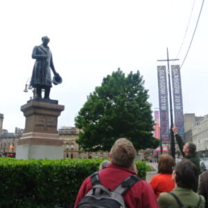 A tour group gathers around the James Oswald statue in Glasgow’s George Square, listening to a guide pointing upward. In the background city buildings line the square.
