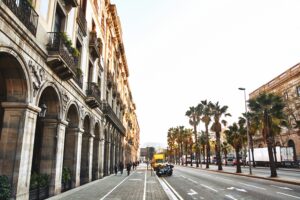 Wide street lined with palm trees and historic buildings featuring arched colonnades, with pedestrians walking and a motorcycle and yellow van driving along designated lanes under a clear sky.