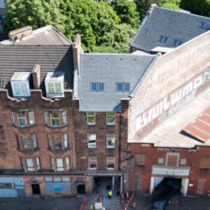 Aerial view of a red sandstone tenement and adjacent industrial building in Glasgow, with visible roof details and faded signage on the gable end.