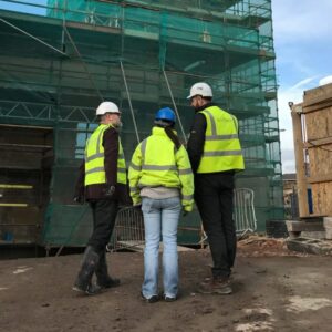 Three people in high-visibility jackets and hard hats stand in front of a historic building surrounded by scaffolding. The scene suggests ongoing restoration or conservation work on the historic pumphouse