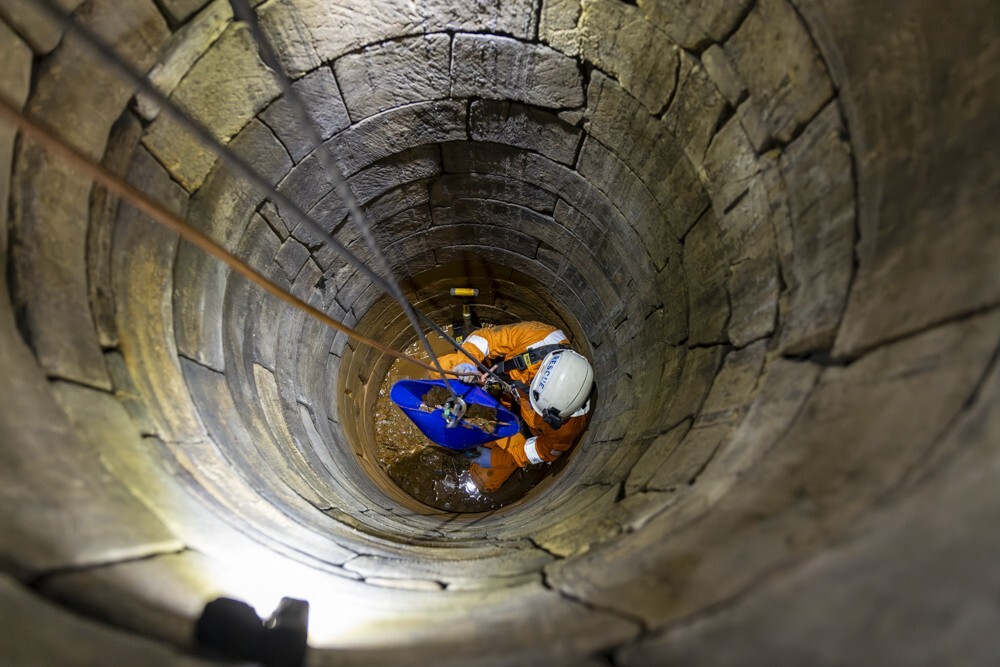 A person in an orange boiler suit wearing a hard had is being hoisted down a stone well.