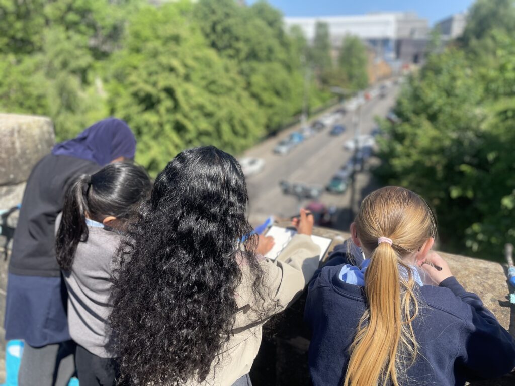 A group of children lean on a wall looking down onto a wall. They are holding pens & paper.