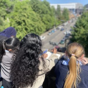 A group of children lean on a wall looking down onto a wall. They are holding pens & paper.