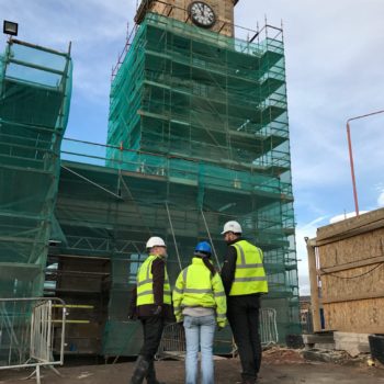 Three people wearing high viz jackets stand in front of a building covered in scaffolding